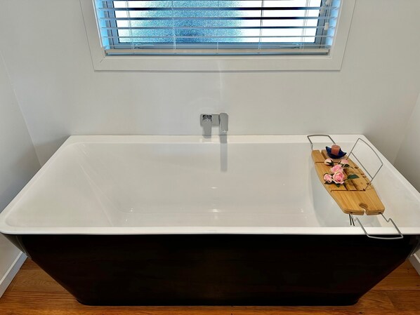 A modern bathroom with a freestanding bathtub, a window with blinds, and a wooden tray with bath items.