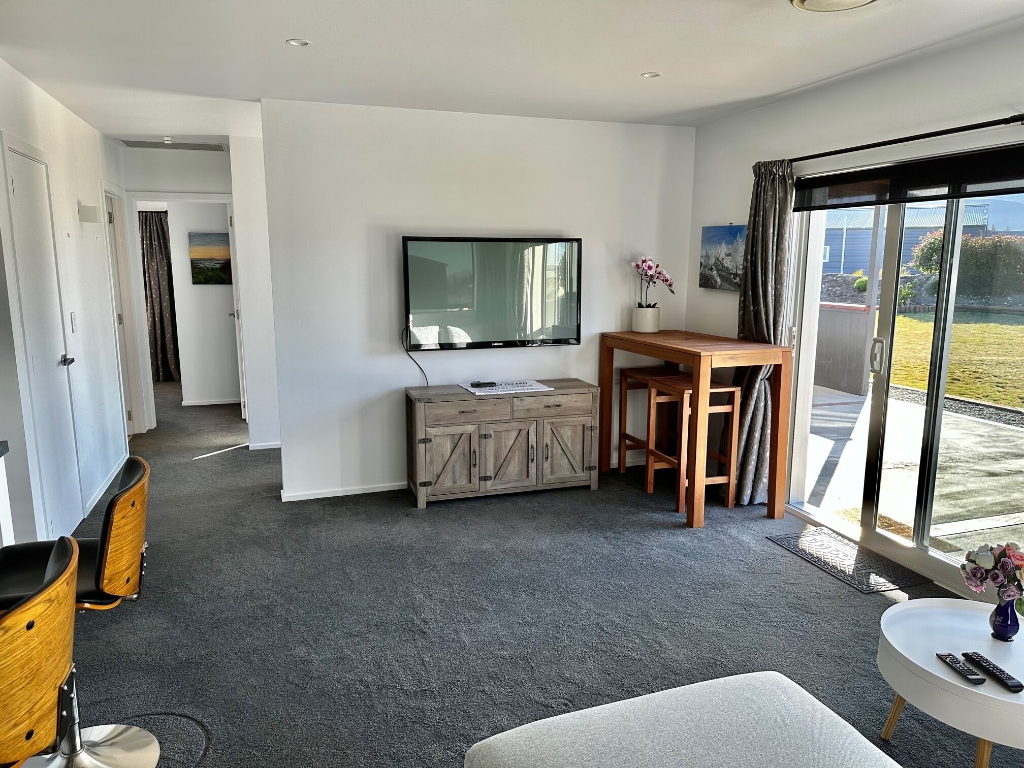 A modern living room with a flat-screen TV, a wooden table, and a sofa. There is a balcony with a view of the outdoors.