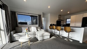 A modern living room with a grey sectional sofa, a round coffee table with a vase of flowers, and a kitchen area with bar stools and a black TV.
