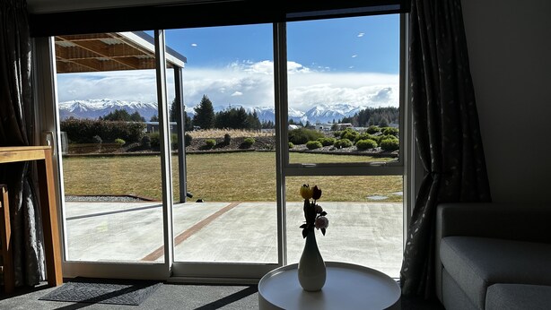 A room with a large sliding glass door leading to a view of snow-capped mountains and a lawn. There is a white vase with flowers on a small round table.