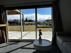 A room with a large sliding glass door leading to a view of snow-capped mountains and a lawn. There is a white vase with flowers on a small round table.