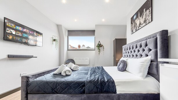 A modern bedroom with a large bed, a mounted TV displaying a streaming service, a cityscape view, and a decorative wall piece.
