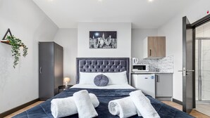 A modern hotel room with a large bed, a cityscape artwork above the bed, a kitchenette, and a shower area.