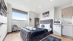 A modern bedroom with a large bed, a small kitchenette, and a window providing a view of the sky.