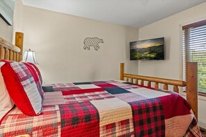 A bedroom with a quilted bedspread, a wooden headboard, a wall-mounted television, and a window with blinds.