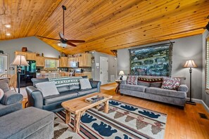 A spacious living area with a wooden ceiling, a large painting, and a kitchen in the background.