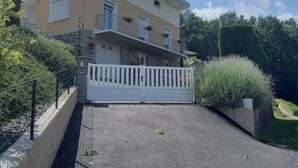 A two-story building with a white fence, a paved driveway, and a garden with various plants. - Appartement en Aveyron (Aubin)