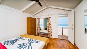 A bedroom with a bed, a wooden wardrobe, a small table, and a view of the beach through an open door. - Dersy Beach Resort (Canacona)