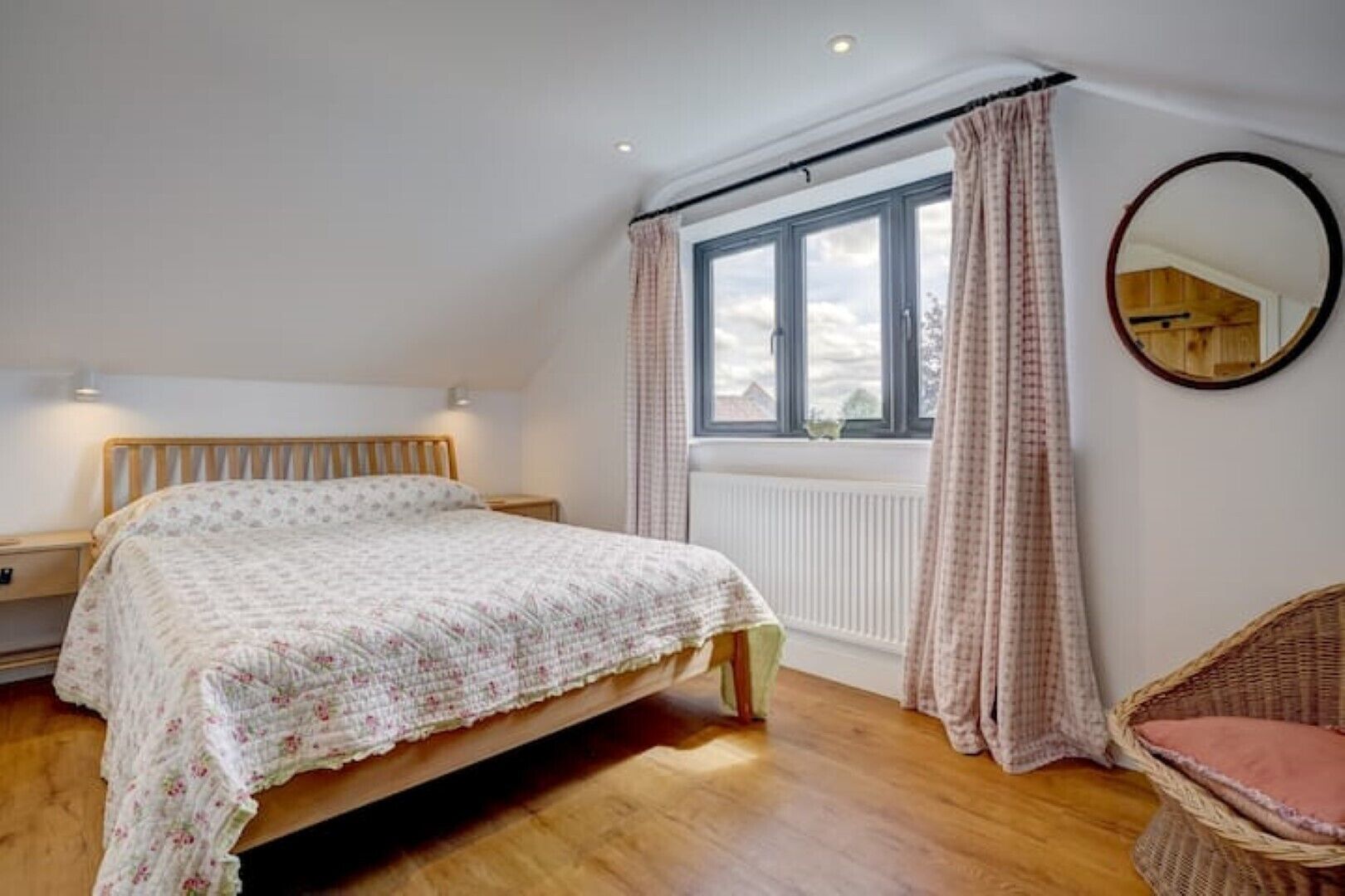 Une chambre à coucher avec un lit, une table de chevet, une chaise en osier, une fenêtre avec des rideaux et un miroir rond accroché au mur.