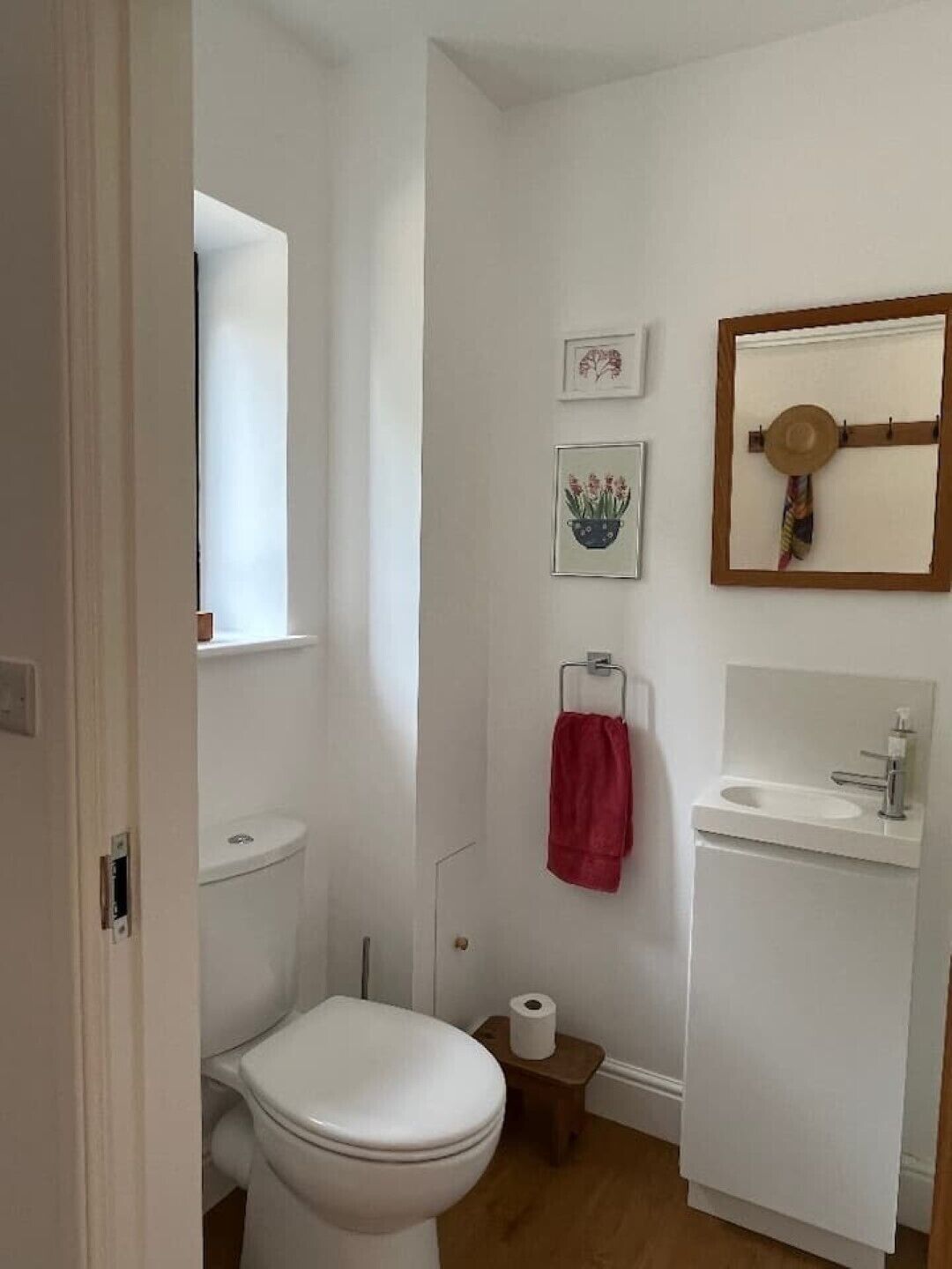 Un baño pequeño y limpio con inodoro, lavamanos y espejo. Hay cuadros enmarcados en la pared y una toalla roja colgada.