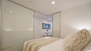 A modern bedroom with a large bed, a glass door closet, a desk with a chair, and a TV mounted on the wall. - Lovely Studio Close to the Wharf (Auckland)
