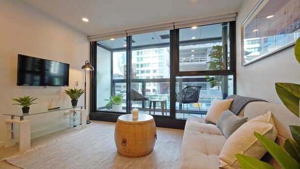 A modern living room with a sofa, a coffee table, a TV, and large windows offering a view of the city. - Lovely Studio Close to the Wharf (Auckland)