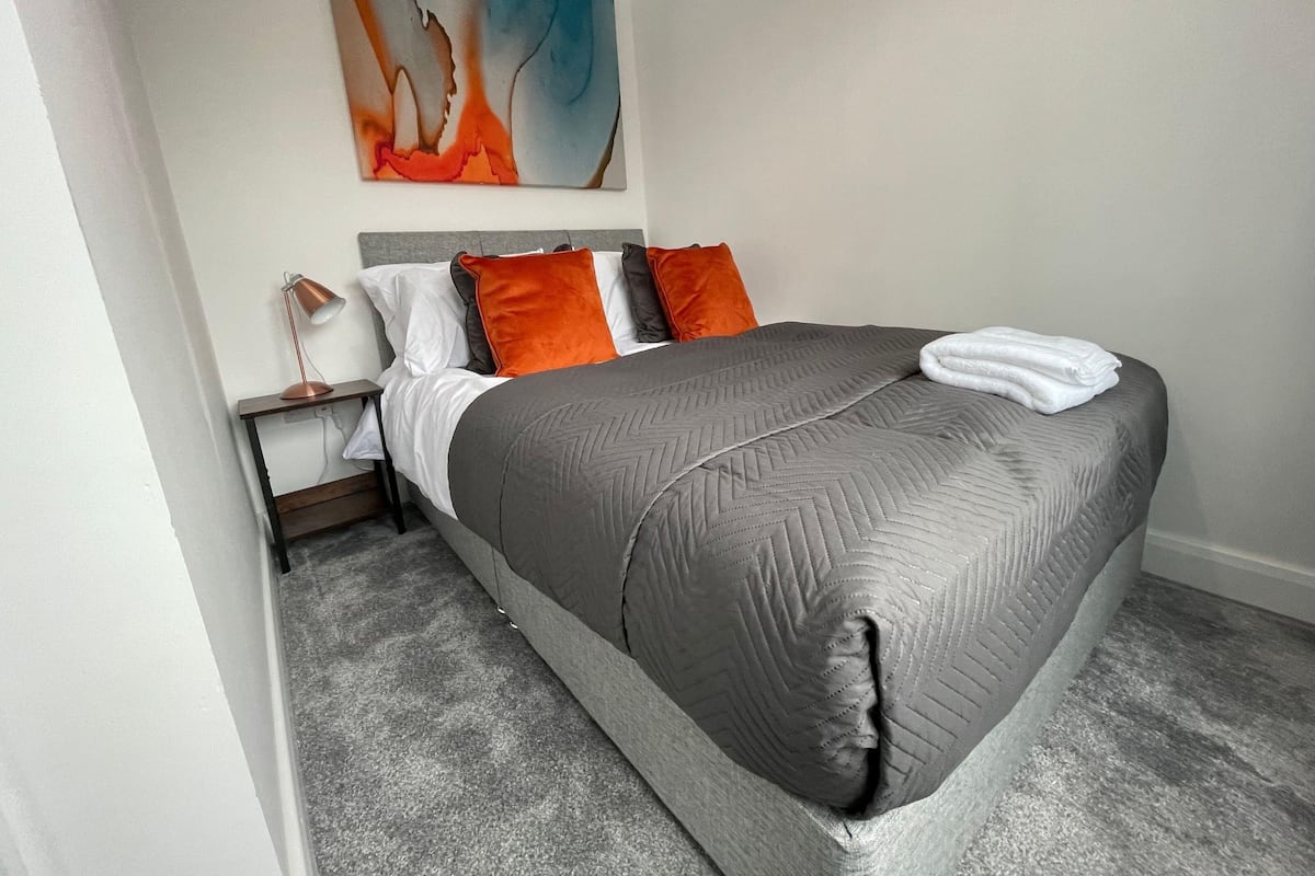 Un dormitorio moderno con una cama gris, almohadas blancas y anaranjadas, un cabecero gris y un cuadro sobre la cama.