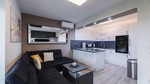 Een moderne woonkamer met een zwarte bank, een klein bijzettafeltje en een keukenhoek met witte kasten en een zwarte magnetron.