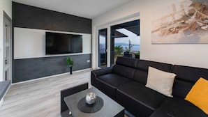 Een moderne woonkamer met een zwarte leren bank, een salontafel met een decoratief glazen object, een flatscreen-televisie aan een donkere muur en een groot abstract schilderij.