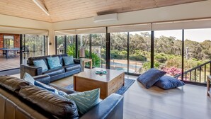 Ein großzügiger Wohnbereich mit einer hohen Holzbalkendecke, einem schwarzen Ledersofa, einem Holztisch und Blick auf Pool und Garten durch große Fenster.