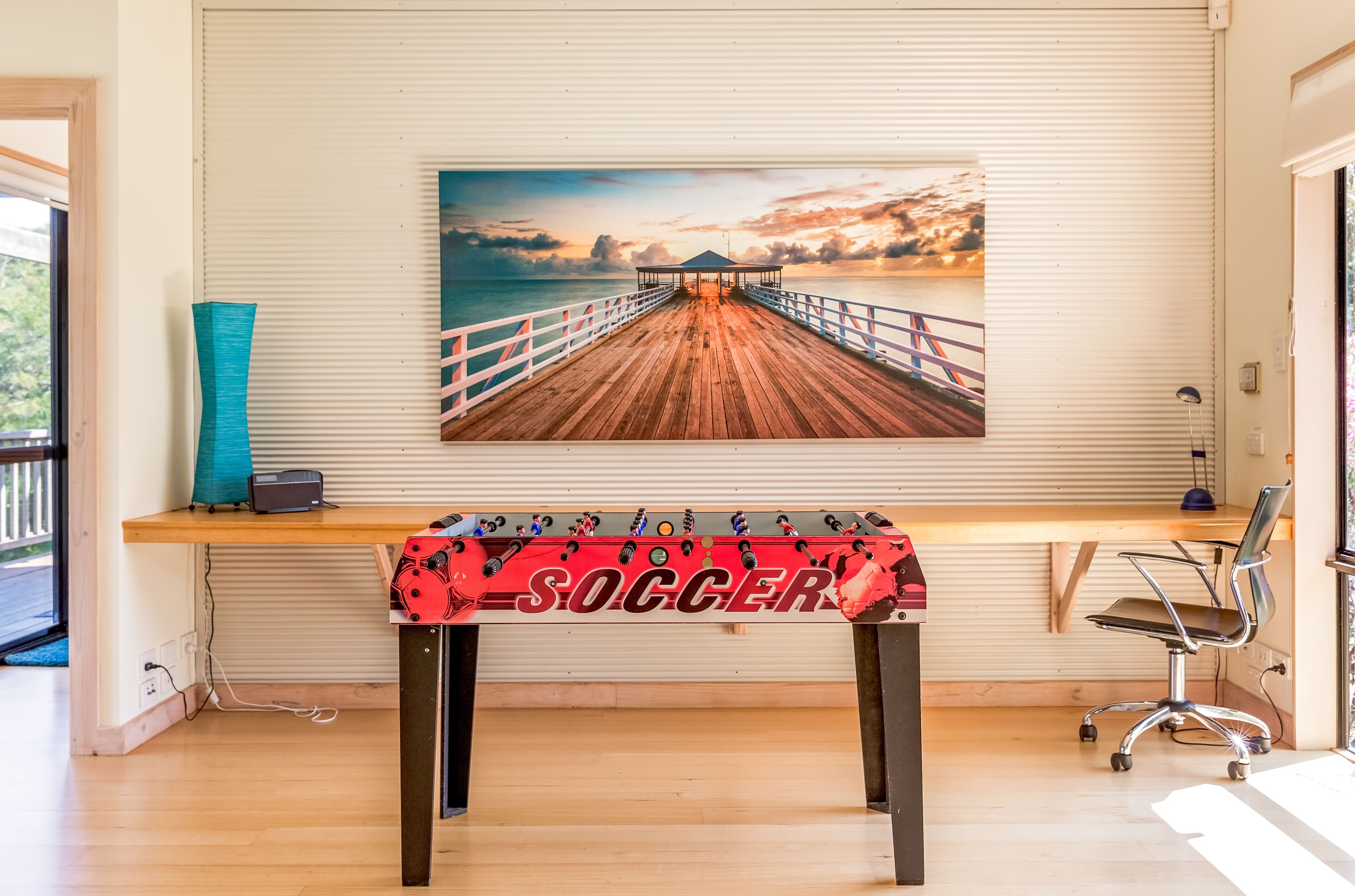 A room with a wooden desk, a soccer-themed tablecloth, a painting of a pier, and a blue vase.
