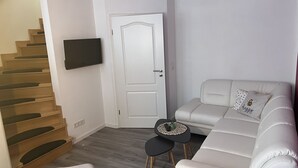 Una sala de estar moderna con una escalera, una puerta blanca, un televisor de pantalla plana montado en la pared, un sofá blanco con un cojín decorativo y una mesita negra con un posavasos tejido.