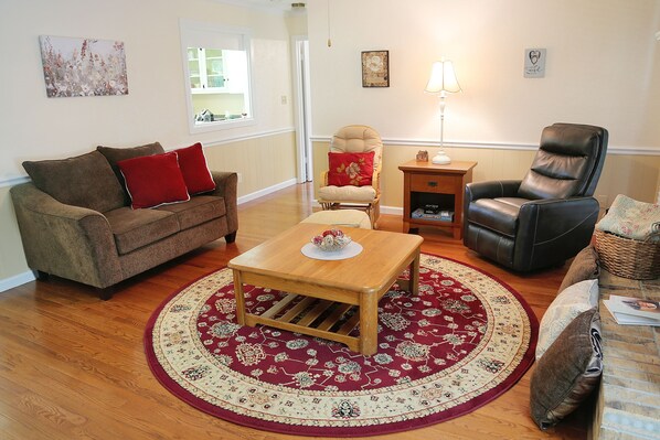 A living room with a brown sofa, a wooden coffee table, a red and beige patterned rug, a leather armchair, and a small side table with a lamp.