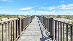Une promenade en bois qui mène à une plage bordée de dunes de sable et sous un ciel bleu dégagé.