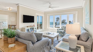 Un salon spacieux doté d’une grande fenêtre offrant une vue sur la mer, d’un ventilateur de plafond et d’un téléviseur à écran plat fixé au mur.