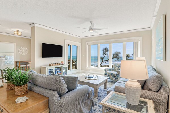 A spacious living room with a large window offering a view of the sea, a ceiling fan, and a flat-screen TV mounted on the wall. - Top Floor Oceanfront 3 Bedroom villa with panoramic views and community pool (Hilton Head Island)
