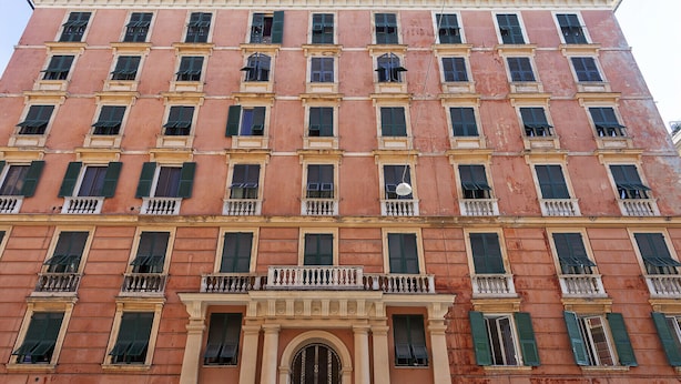 Un immeuble à plusieurs étages doté d’une façade symétrique, avec des fenêtres régulièrement espacées et des volets verts, une entrée centrale surmontée d’un portail en arc de cercle et un balcon agrémenté de balustrades.
