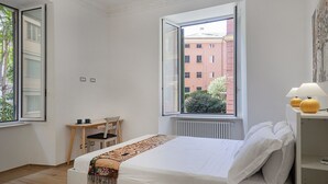 Une chambre à coucher comprenant un lit, une chaise, une table avec une lampe et une fenêtre donnant sur des immeubles.