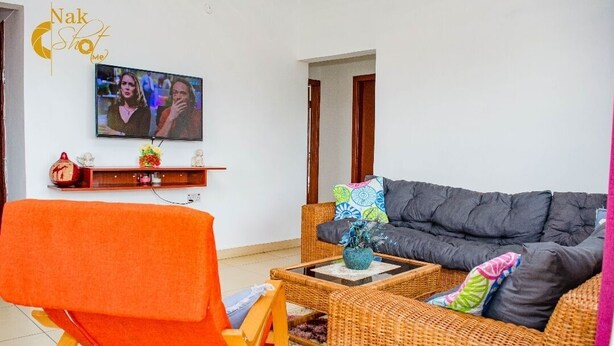 A living room with a television, wicker furniture, and a wall-mounted shelf. - 2 Bedroom Cozy Apartment. (Accra)