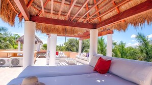 Un área de estar exterior techada con sofás blancos y un cojín rojo, rodeada de abundante vegetación y una estructura de techo de paja.