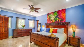 A bedroom with a wooden bed, a ceiling fan, a large painting of a frog, and a nightstand with a lamp.
