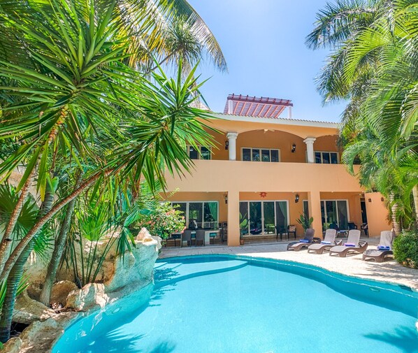 A two-story building with a swimming pool, surrounded by palm trees and a red structure in the background.