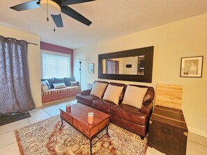 A living room with a brown leather sofa, a wooden coffee table, a patterned rug, a ceiling fan, and a large mirror on the wall.