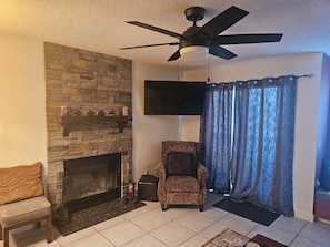 A living room with a stone fireplace, a television mounted on the wall, a patterned armchair, and a ceiling fan. - 4314 Mountain View Suite w/Pool, hot tub & more (Beech Mountain)