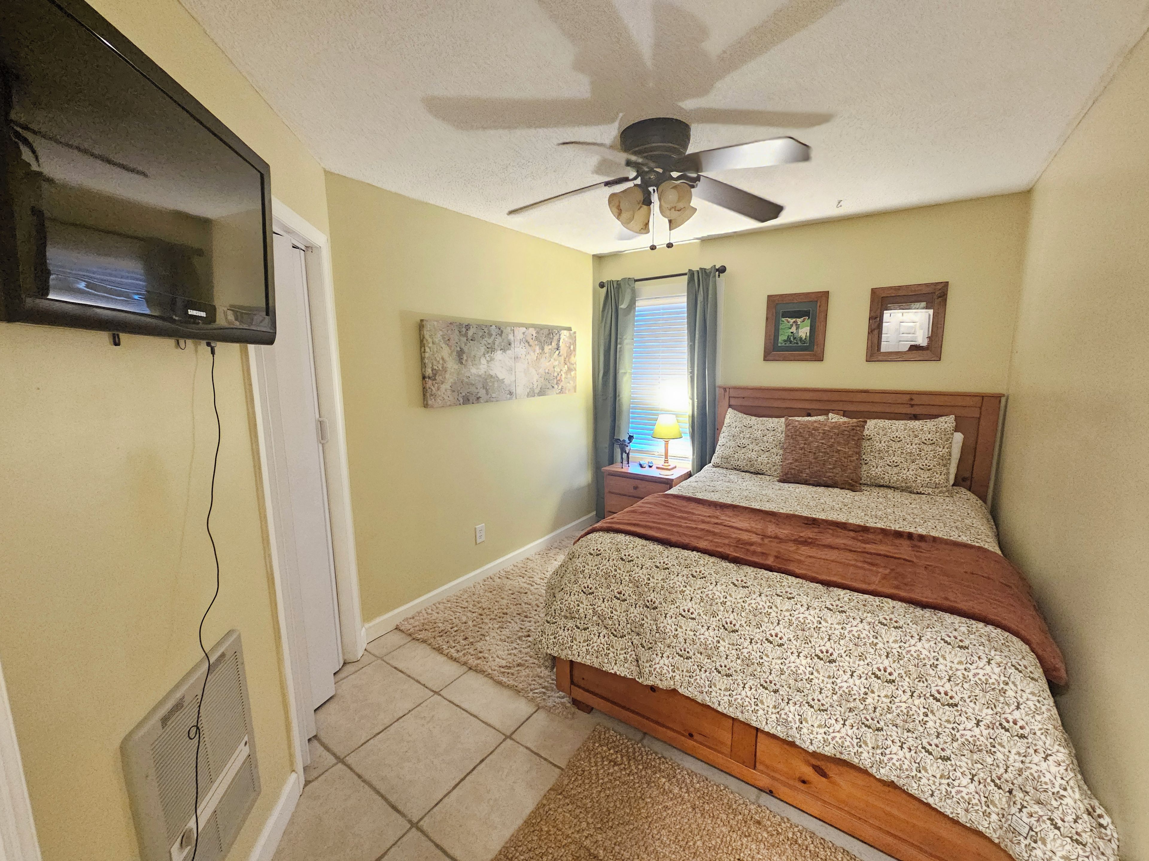 Une chambre à coucher comprenant un lit, un ventilateur de plafond, des œuvres d’art et un téléviseur fixé au mur.