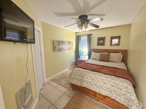 Une chambre à coucher comprenant un lit, un ventilateur de plafond, des œuvres d’art et un téléviseur fixé au mur.