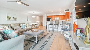Ein modernes Wohnzimmer mit einer Couch, einem Hocker und einem Essbereich mit Stühlen. Im Hintergrund ist eine Küche mit Holzschränken und einem schwarzen Kühlschrank zu sehen.