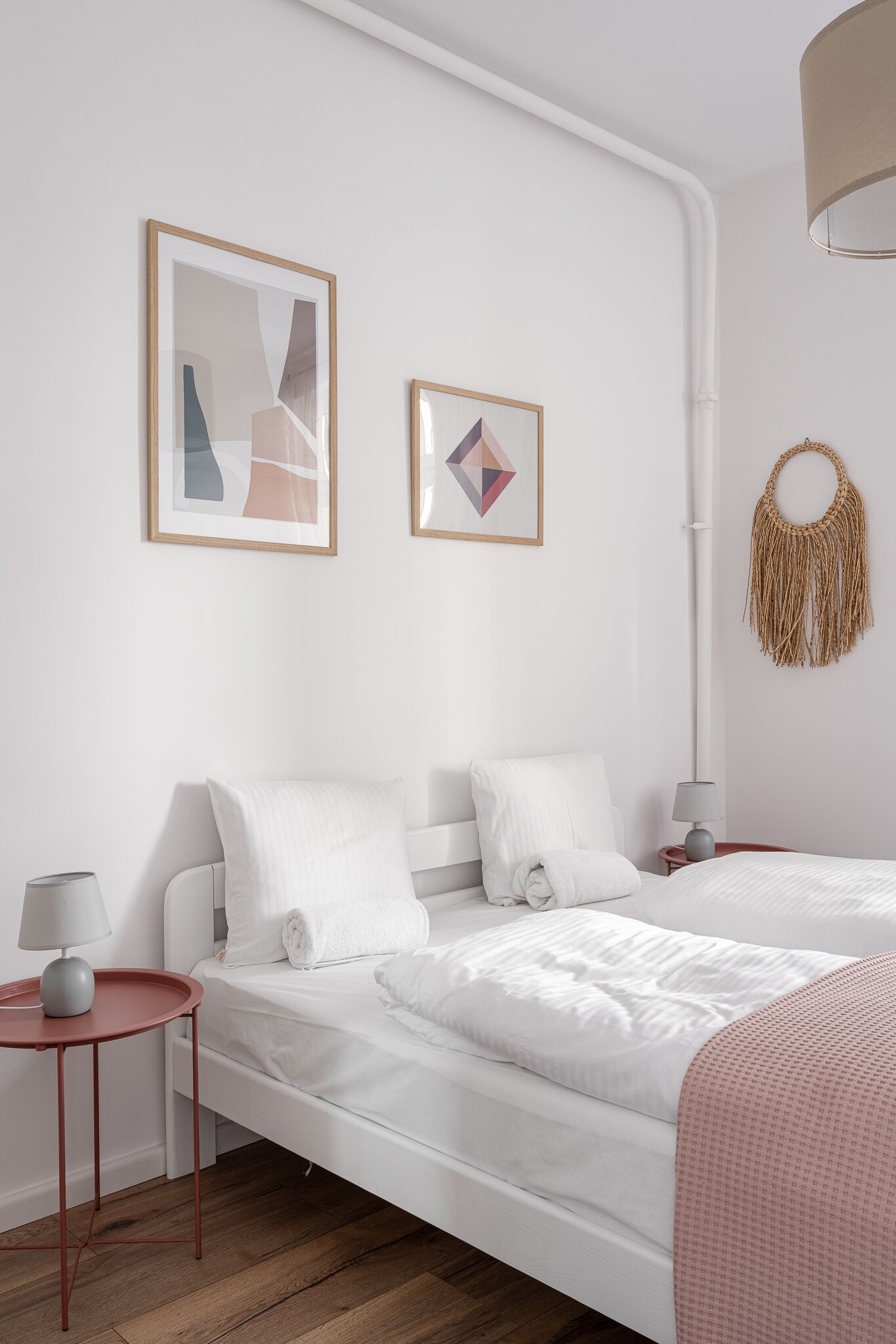 Et moderne soveværelse med en hvid seng, to indrammede abstrakte malerier, et lille sengebord med en lampe og en vægophæng i makramé.