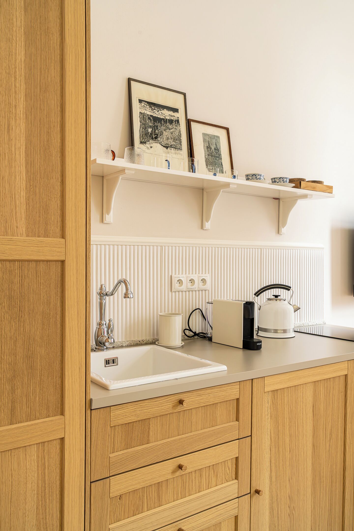A kitchen with wooden cabinets, a white countertop, a sink, a kettle, and framed pictures on a shelf.