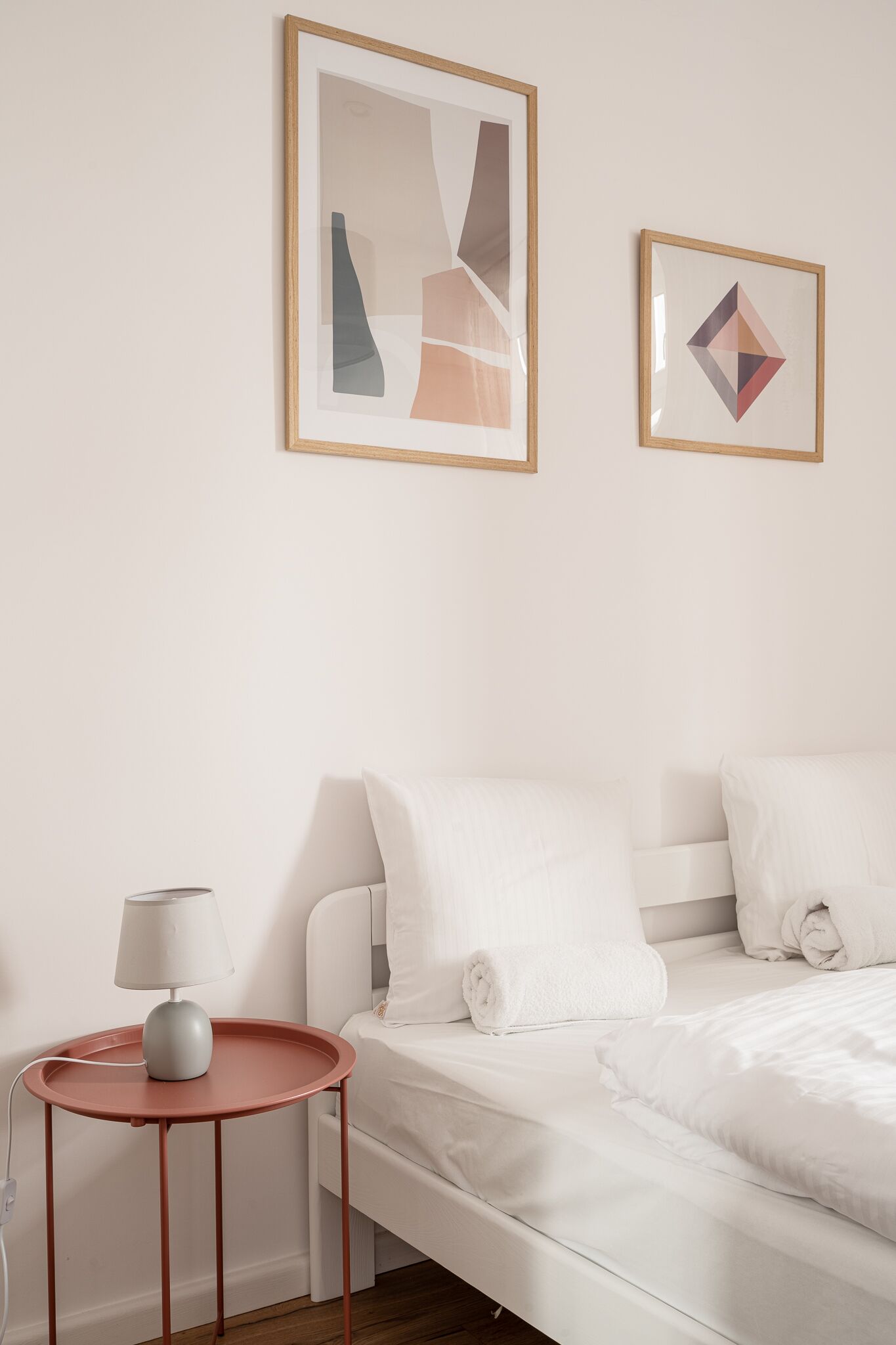 A neatly arranged bedroom with a bed, a bedside table with a lamp, and two framed abstract artworks on the wall.