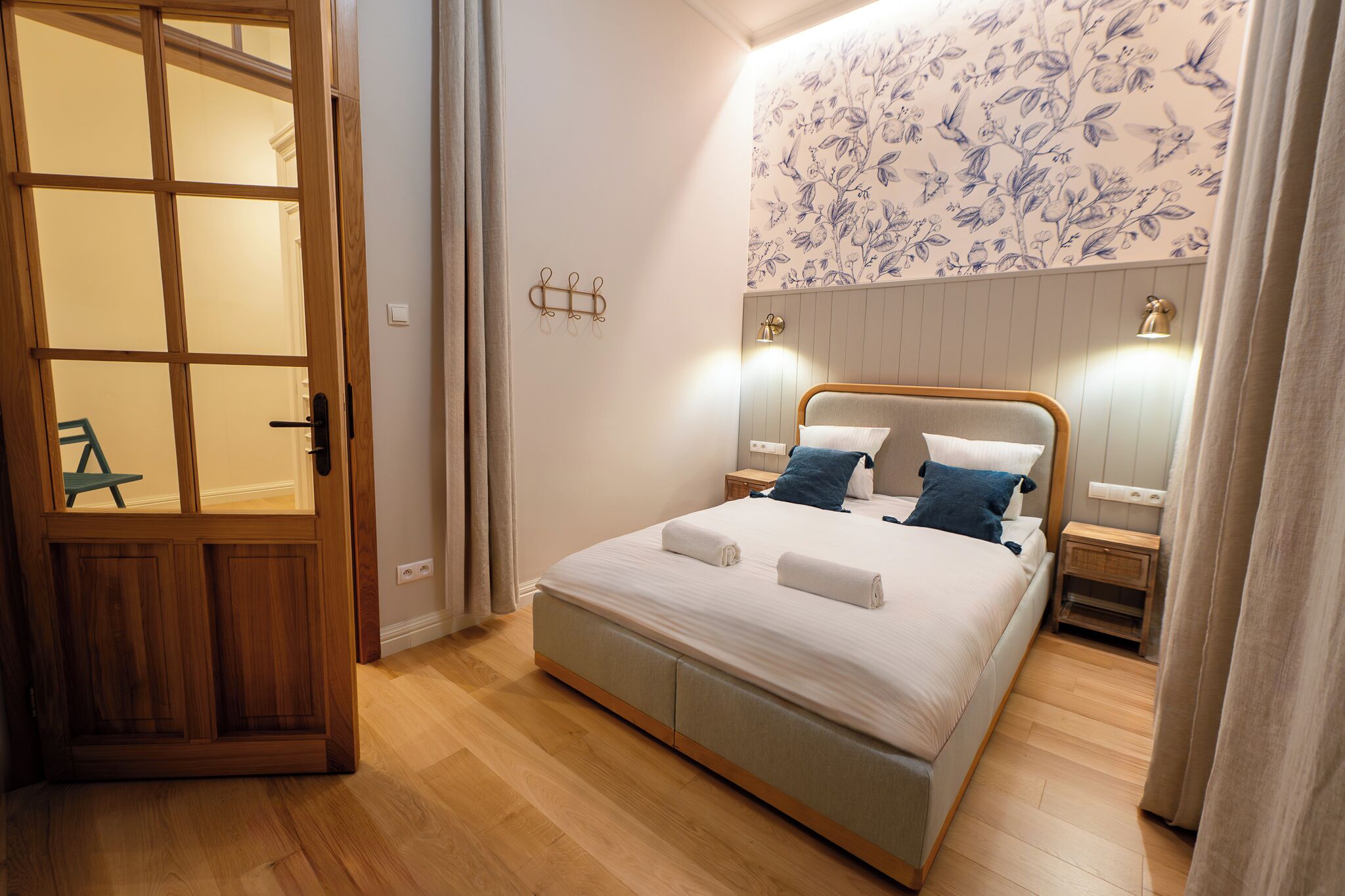 A bedroom with a wooden door, a bed with two pillows, a nightstand, and a wall-mounted light fixture.