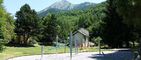 A playground with climbing structures and a small building, surrounded by trees and a mountain in the background.