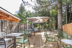 An outdoor patio with wooden flooring, green metal chairs, and tables, surrounded by trees and a wooden fence.