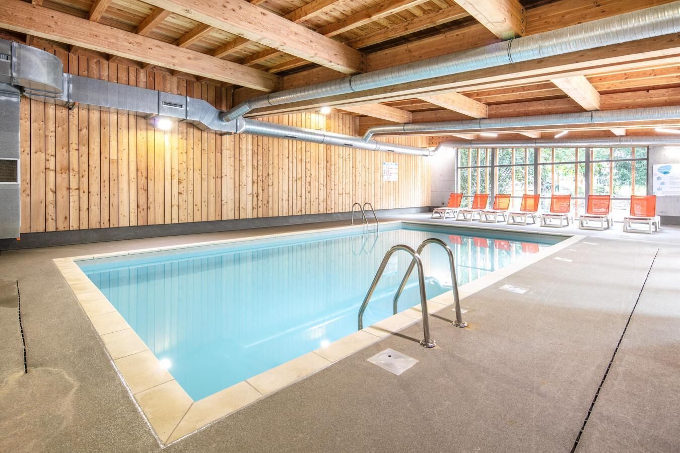 Une piscine intérieure aux murs recouverts de panneaux de bois, avec un sol en béton et une rangée de chaises orange le long du bord.