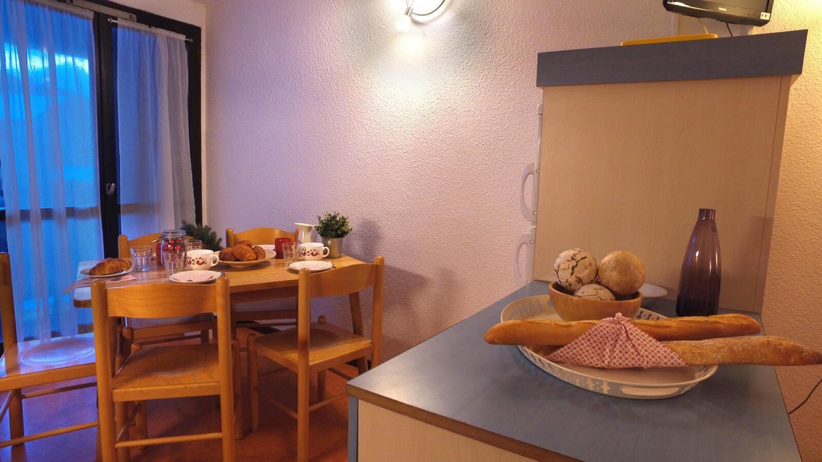 A cozy dining area with a wooden table set for breakfast, including croissants, a croissant-shaped cake, and a bowl of cookies.