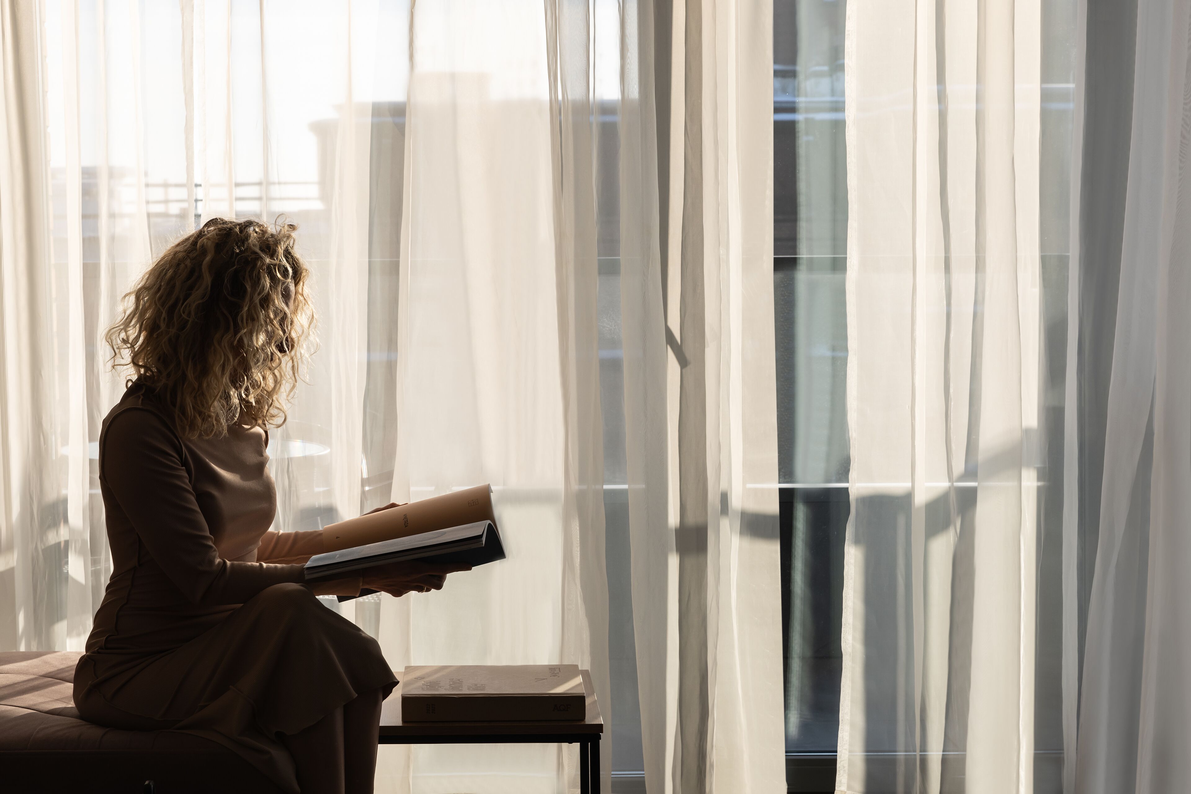Une personne assise près d’une fenêtre, derrière des rideaux légers, est en train de lire un livre.