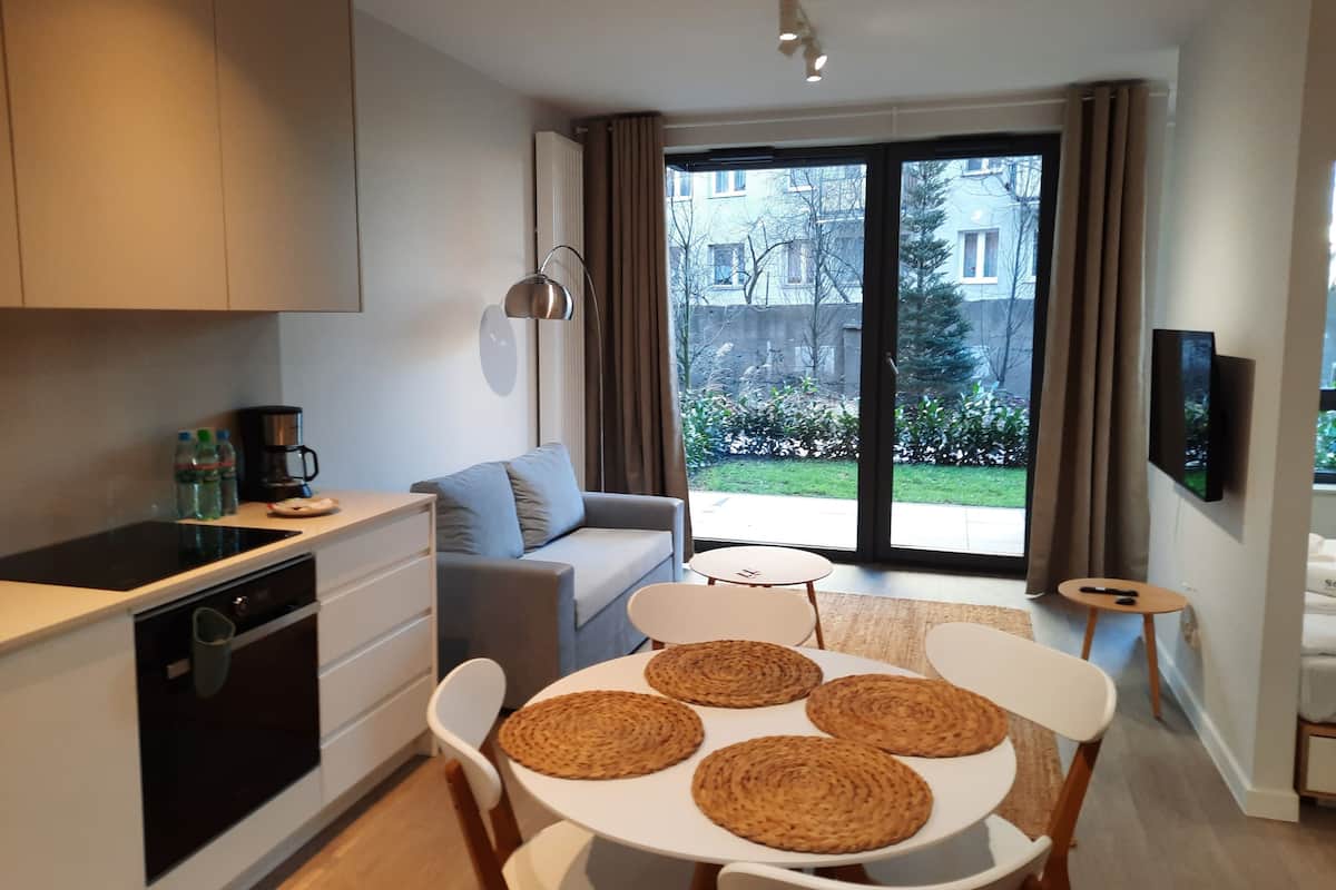 A modern kitchen with a built-in oven, a dining area with a round table and woven placemats, a grey sofa, and a sliding glass door leading to a balcony with a view of buildings and greenery.