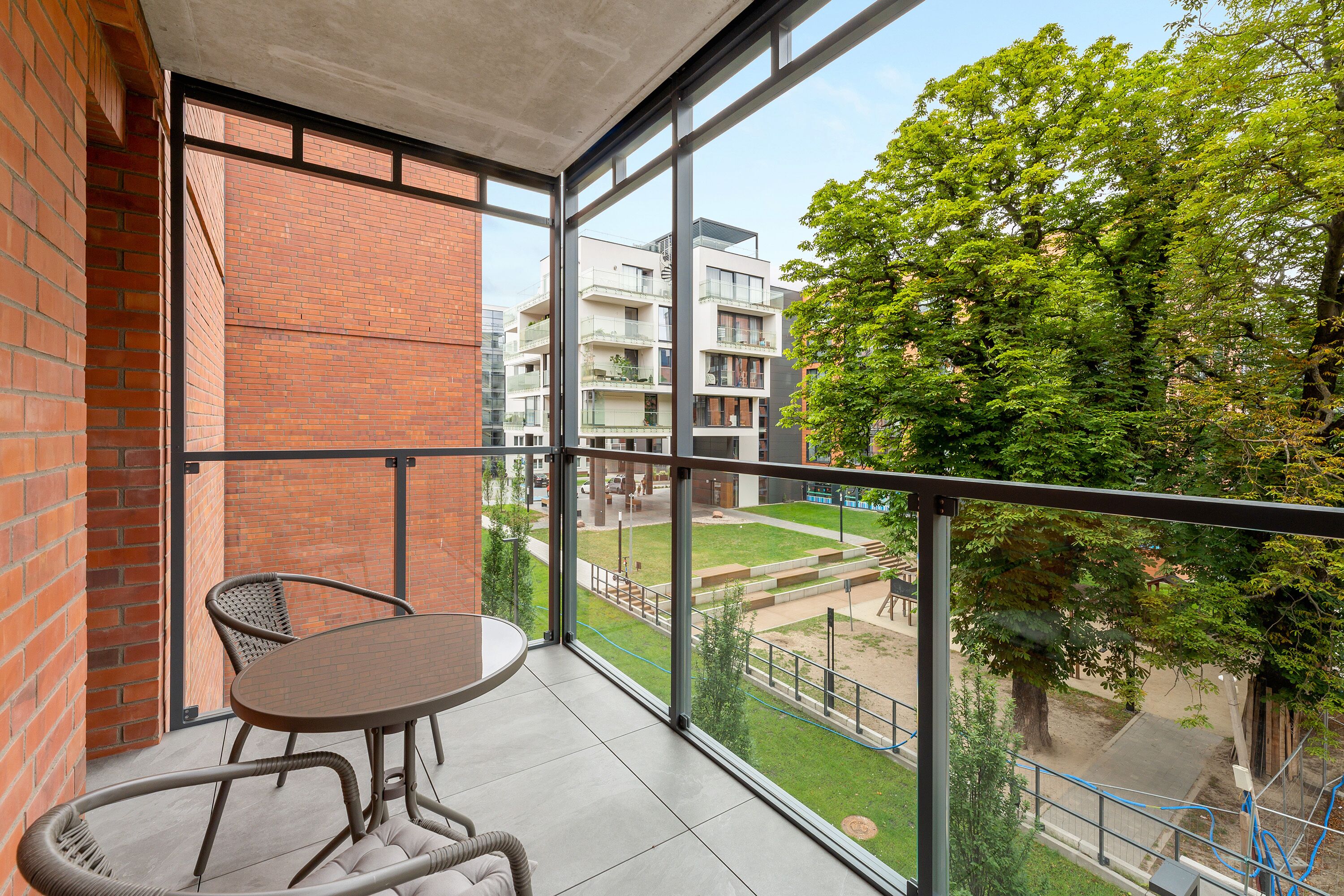 A balcony with a table and chairs, a view of a courtyard with buildings and trees.