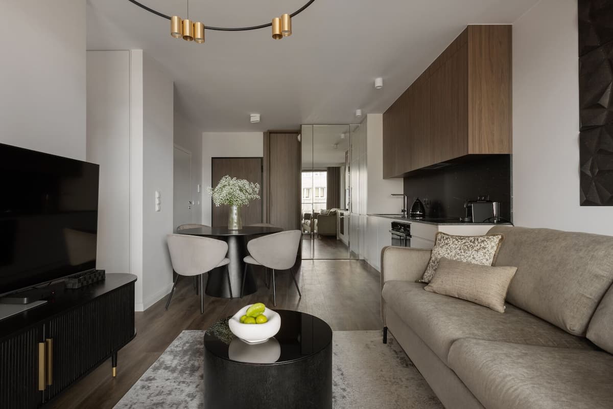 A modern living room with a flat-screen TV, a sofa, a dining table with chairs, and a kitchen area with wooden cabinets and a black countertop.