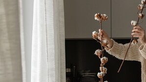 Une personne arrange une branche de coton dans un vase noir posé sur une table en bois.